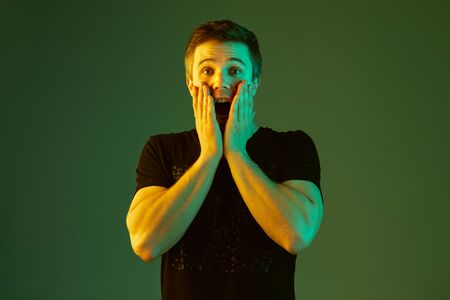 Shocked, hands on head. Caucasian mans portrait isolated on green studio background in neon light. Beautiful male model in black shirt. Concept of human emotions, facial expression, sales, ad.の写真素材