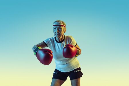Still kicking. Senior woman in sportwear boxing on gradient background, neon light. Female model in...