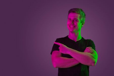 Smiling, hands crossed, pointing at side. Caucasian man's portrait on purple studio background in...