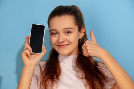 Showing phone screen, thumb up. Caucasian teen girls portrait on blue
