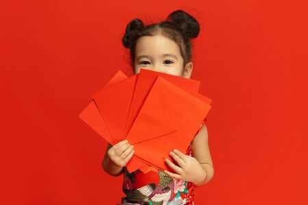 Holding red envelopes, looks happy, smiling. Happy Chinese New Year 2020. Asian cute little girl isolated on red background in traditional clothing. Celebration, human emotions, holidays. Copyspace.の写真素材