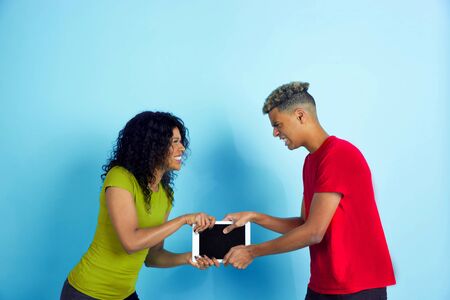 Fight for tablet for fun. Young emotional african-american man and woman in colorful casual clothes on blue background. Beautiful couple. Concept of human emotions, facial expession, relations, ad.の写真素材
