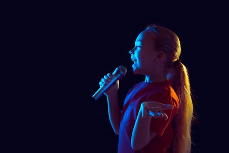 Singing happy. Caucasian girls portrait on dark studio background in neon light. Beautiful female model with speaker. Concept of human emotions, facial expression, sales, ad, hobby, dream, music.の写真素材