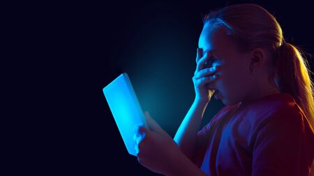 Shocked, scared. Caucasian girl's portrait on dark background in neon light. Beautiful female model using tablet. Concept of human emotions, facial expression, sales, ad, modern tech, gadgets. Flyer.の写真素材
