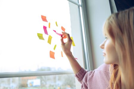 Beautiful caucasian business lady working in office. Young female model in co-working place makes notes on stickers, while sitting near by window. Concept of business, finance, freelance, open space.の写真素材