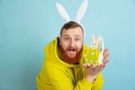 Holding traditional decor. Caucasian man as an Easter bunny with bright casual clothes on blue...
