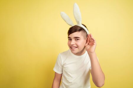 Listen to secret. Caucasian boy as an Easter bunny on yellow studio background. Happy easter greetings. Beautiful male model. Concept of human emotions, facial expression, holidays. Copyspace.の写真素材