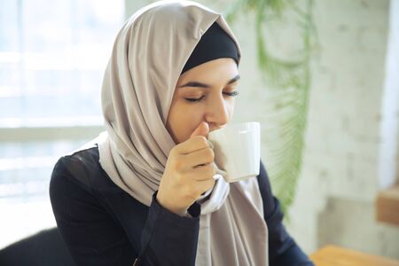 Enjoying coffee. Portrait of a beautiful arabian businesswoman wearing hijab while working at openspace or office. Concept of occupation, freedom in business area, leadership, success, modern solution.の写真素材