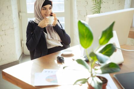 Talking on phone while drinking coffee. Portrait of a beautiful arabian businesswoman wearing hijab while working at openspace or office. Concept of occupation, leadership, success, modern solution.の写真素材
