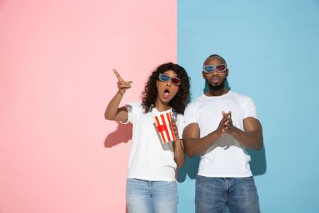 Watching cinema, shocked. Young man and woman in casual clothes on pink, blue bicolored background....
