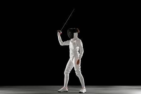 Graceful. Teen girl in fencing costume with sword in hand isolated on black background. Young female...