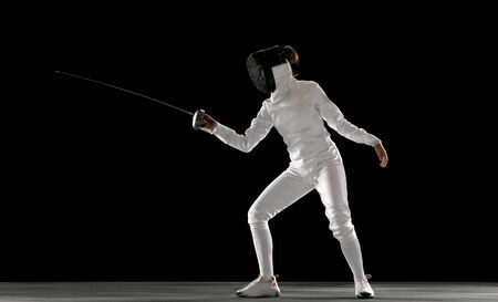 Invincible. Teen girl in fencing costume with sword in hand isolated on black background. Young...
