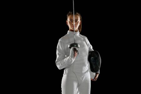 Confident. Teen girl in fencing costume with sword in hand isolated on black background. Young female model practicing and training in motion, action. Copyspace. Sport, youth, healthy lifestyle.の写真素材