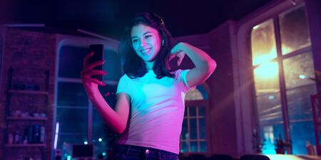 Taking selfie, vlog. Cinematic portrait of handsome stylish woman in neon lighted interior. Toned...