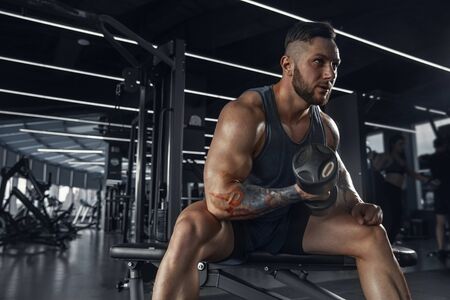 Confident. Young muscular caucasian athlete practicing in gym with the weights. Male model doing...