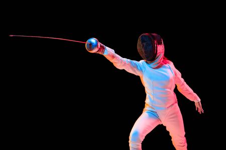 Attacking. Teen girl in fencing costume with sword in hand isolated on black background, neon light....