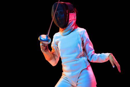 Close up. Teen girl in fencing costume with sword in hand isolated on black background, neon light. Young model practicing and training in motion, action. Copyspace. Sport, youth, healthy lifestyle.の写真素材