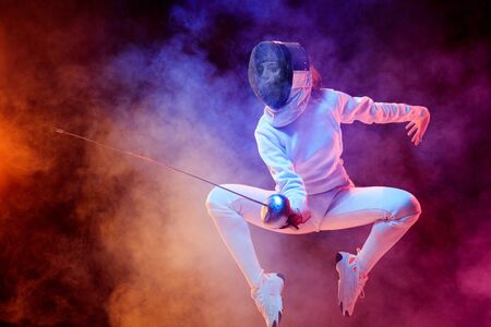 Blooming. Teen girl in fencing costume with sword in hand isolated on black background, neon lighted smoke. Practicing and training in motion, action. Copyspace. Sport, youth, healthy lifestyle.の写真素材