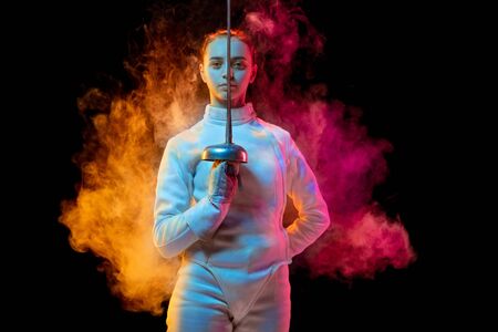 Historical time. Teen girl in fencing costume with sword in hand isolated on black background, neon lighted smoke. Practicing and training in motion, action. Copyspace. Sport, youth, healthy lifestyle.の写真素材