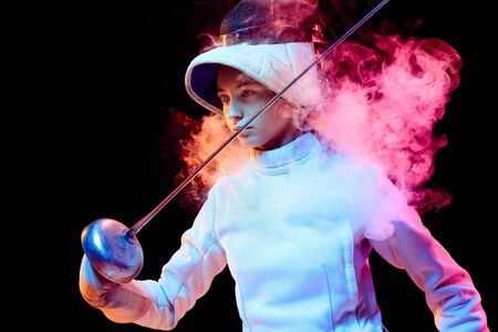 Pride. Teen girl in fencing costume with sword in hand isolated on black background, neon lighted smoke. Practicing and training in motion, action. Copyspace. Sport, youth, healthy lifestyle.の写真素材