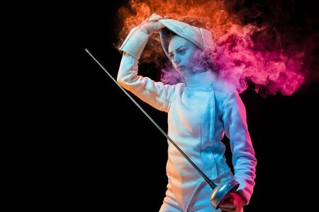 Winner thoughts. Teen girl in fencing costume with sword in hand isolated on black background, neon lighted smoke. Practicing and training in motion, action. Copyspace. Sport, youth, healthy lifestyle.の写真素材
