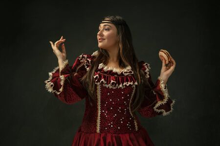 First love. Portrait of medieval young woman in red vintage clothing holding burger on dark background. Female model as a duchess, royal person. Concept of comparison of eras, modern, fashion, beauty.の写真素材