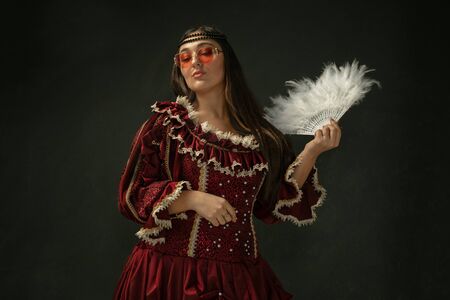 Red eyewear and fluffy fan. Portrait of medieval young woman in red vintage clothing on dark...