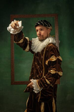 Selfie time. Portrait of medieval young man in vintage clothing with wooden frame on dark background. Male model as a duke, prince, royal person. Concept of comparison of eras, modern, fashion.の写真素材