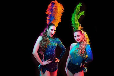 Friends. Beautiful young women in carnival, stylish masquerade costume with feathers on black background in neon light. Copyspace for ad. Holidays celebration, dancing, fashion. Festive time, party.の写真素材