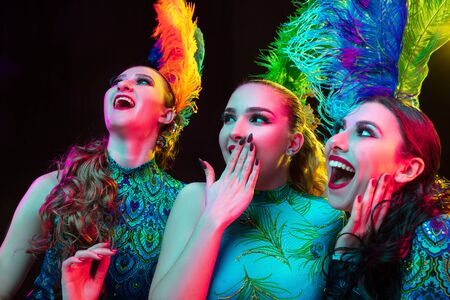 Close up. Beautiful young women in carnival, stylish masquerade costume with feathers on black background in neon light. Copyspace for ad. Holidays celebration, dancing, fashion. Festive time, party.の写真素材