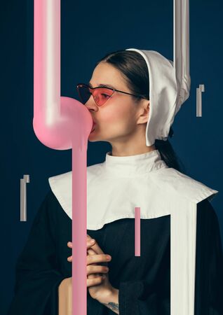 Medieval young woman in black vintage clothing as a nun standing on dark blue background. Trying bubble gum. Concept of comparison of eras. Stylish creative design, art vision, new look of artwork.の写真素材