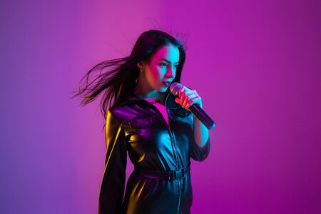 Caucasian female singer portrait isolated on purple studio background in neon light. Beautiful female model in black wear with microphone. Concept of human emotions, facial expression, ad, music, art.の写真素材