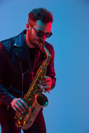 Young caucasian jazz musician playing the saxophone on gradient blue-purple studio background in neon light. Concept of music, hobby, festival. Joyful attractive guy. Colorful portrait of artist.の写真素材