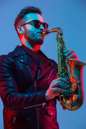Young caucasian jazz musician playing the saxophone on gradient blue-purple studio background in neon light. Concept of music, hobby, festival. Joyful attractive guy. Colorful portrait of artist.の写真素材