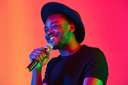 Young african-american musician singing on gradient orange-purple studio background in neon light. Concept of music, hobby, festival. Joyful party host, stand upper. Colorful portrait of artist.の写真素材