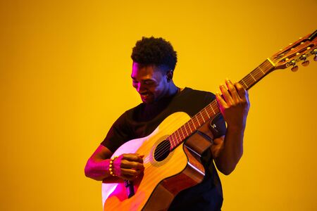 Young and joyful african-american musician playing guitar and singing on gradient orange-yellow...