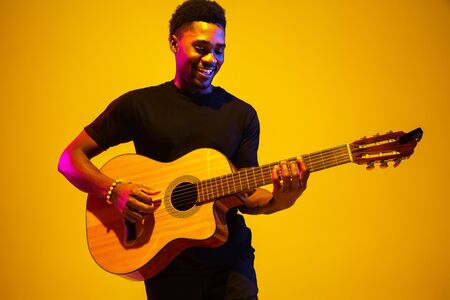 Young and joyful african-american musician playing guitar and singing on gradient orange-yellow...