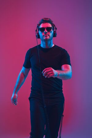 Young caucasian musician in headphones singing on gradient pink-purple background in neon light. Concept of music, hobby, festival. Joyful party host, DJ, stand upper. Colorful portrait of artist.の写真素材