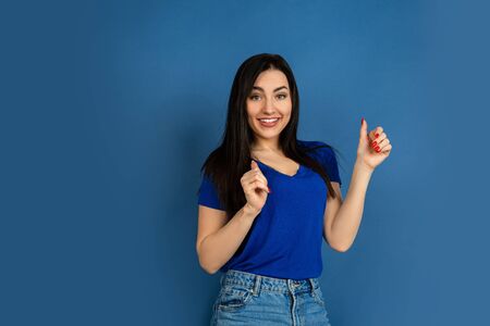 Thumb up. Beautiful caucasian womans portrait isolated on blue studio background. Beautiful female brunette model in casual style. Concept of human emotions, facial expression, sales, ad, copyspace.の写真素材