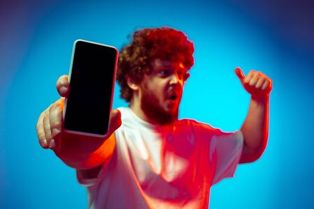 Shows blank screen phone. Caucasian mans portrait isolated on blue studio background in neon light....