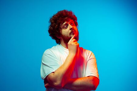 Thoughtful, has an idea. Caucasian mans portrait isolated on blue studio background in neon light. Beautiful male model, red curly hair. Concept of human emotions, facial expression, sales, ad.の写真素材