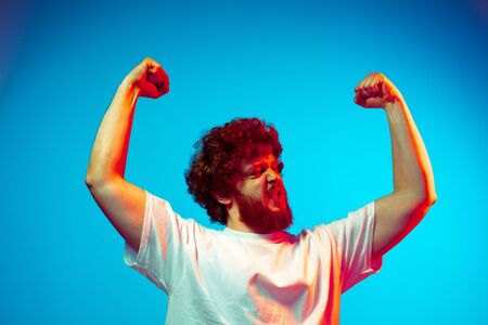 Winning, crazy happy. Caucasian mans portrait isolated on blue studio background in neon light. Beautiful male model, red curly hair. Concept of human emotions, facial expression, sales, ad.の写真素材