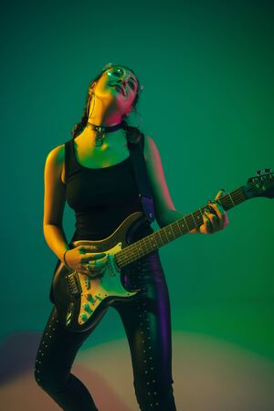 Rockstar. Caucasian female guitarist portrait on green studio background in neon light. Beautiful female model in black wear with guitar. Concept of human emotions, facial expression, ad, music, art.の写真素材
