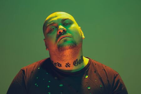 Portrait of a hipster guy with colorful neon light on green studio background. Male model with calm and serious mood. Facial expression, cyberpunk, millenials lifestyle and look like. Fashion.の写真素材