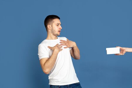 Getting a giftbox. Caucasian young mans portrait on blue studio background. Beautiful male model in casual style, pastel colors. Concept of human emotions, facial expression, sales, ad, celebration.の写真素材