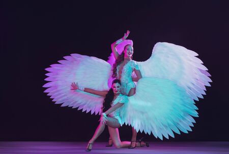 Young female dancers with white angels wings in purple blue neon light on black background. Graceful models, womens dancing, posing. Concept of carnival, beauty, motion, overcoming, blooming.の写真素材