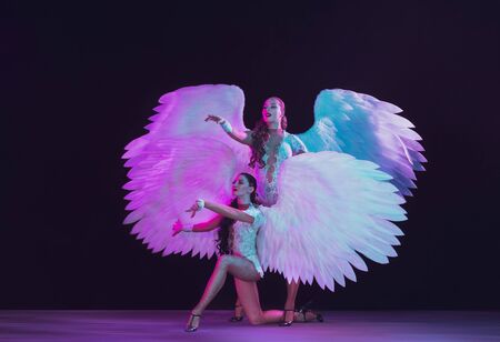 Young female dancers with white angels wings in purple blue neon light on black background. Graceful models, womens dancing, posing. Concept of carnival, beauty, motion, overcoming, blooming.の写真素材
