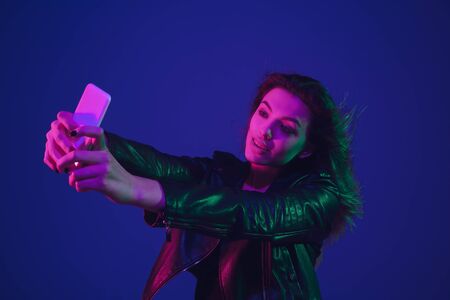 Taking selfie. Caucasian womans portrait isolated on blue studio background in neon light. Beautiful...