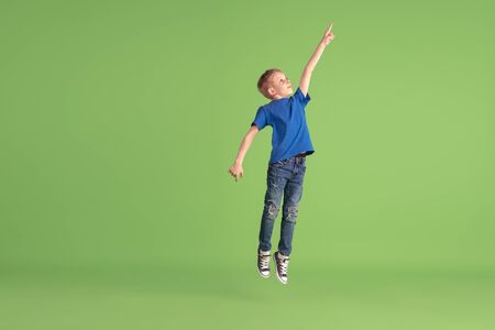 Pointing. Happy boy playing and having fun on green studio background. Caucasian kid in bright cloth looks playful, laughting, smiling. Concept of education, childhood, emotions, facial expression.の写真素材