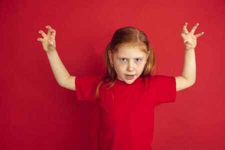 Scary and horrible. Caucasian little girl portrait isolated on red studio background. Cute redhair...